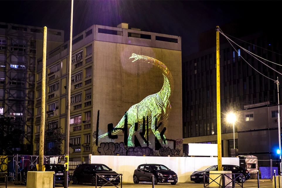 Projection monumentale d’un dinosaure sur une façade d’immeuble – Prehistoric Safari, art urbain préhistorique
