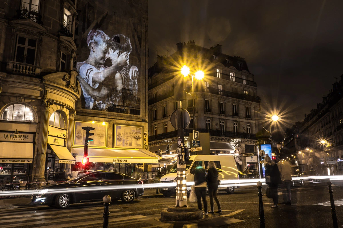 Projection d’un couple s’embrassant sur une façade d’angle parisienne avec circulation nocturne – Le Baiser, art urbain